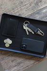 Detailaufnahme einer großen Ablageschale aus Leder in Schwarz auf einer Holzplatte und in dieser befindet sich ein Handy, Kleingeld, ein Kartenetui und ein Schlüsselanhänger.