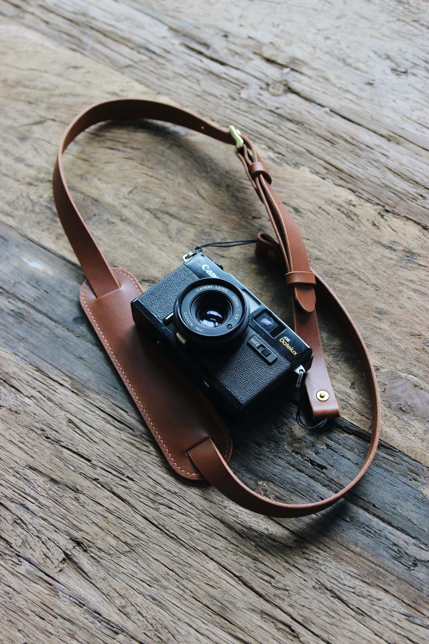 Eine Kamera mit einem Leder Kameragurt in Braun auf einem Holztisch liegend.