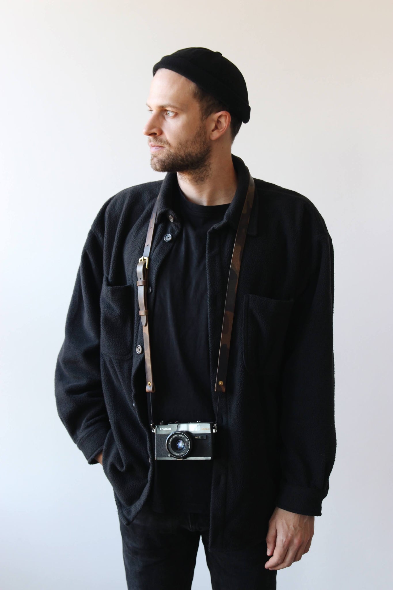 Ein Mann mit schwarzer Kleidung trägt eine Kamera mit einem Leder Kameragurt in Camo um den Hals.