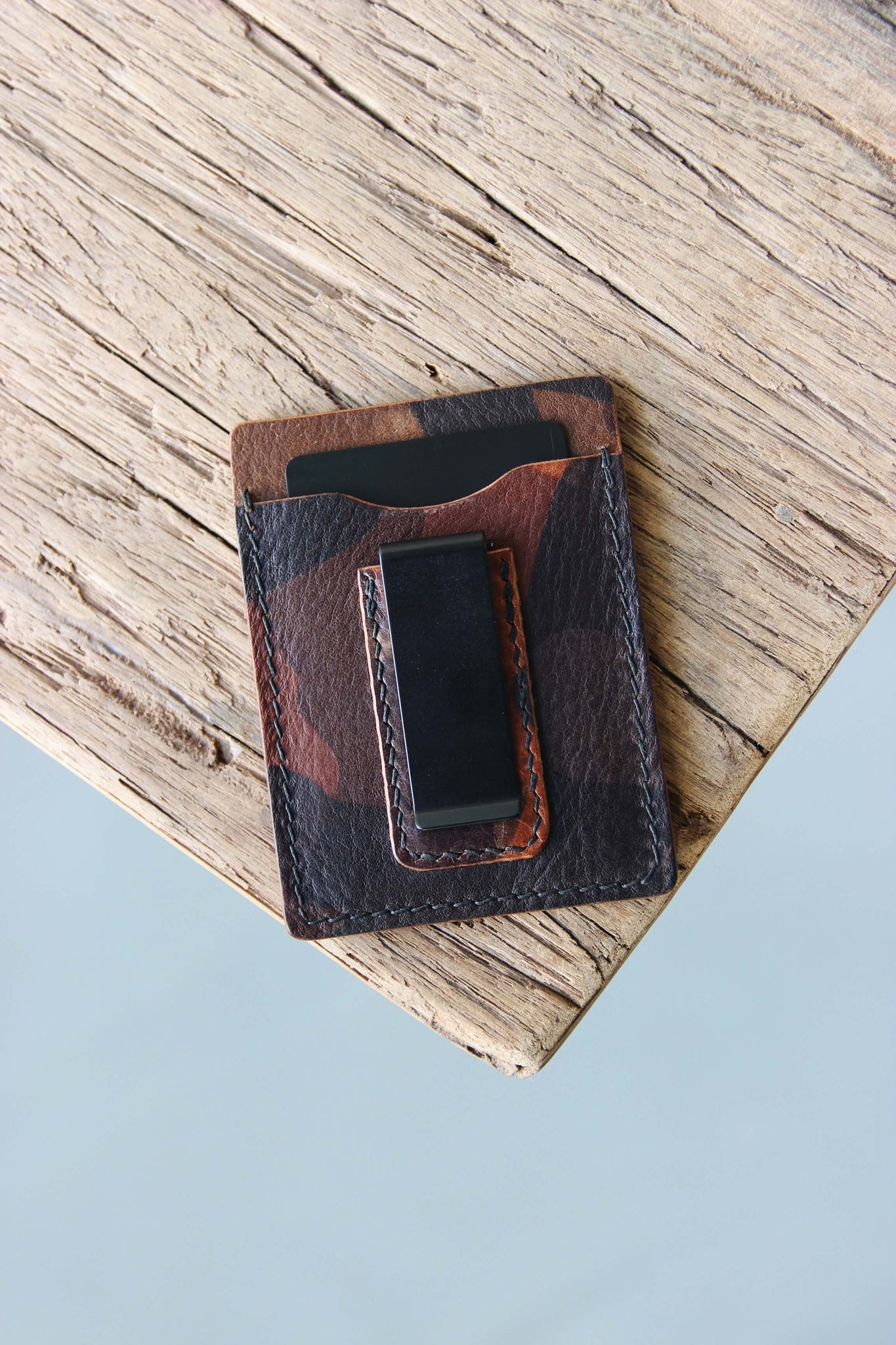Ein Kreditkartenetui aus Leder in Camouflage mit integrierter Geldklammer liegt auf einer Holzplatte.