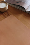 Lederkante einer Schreibtischunterlage aus Leder in Natural auf einer Holzplatte und eine Kaffeetasse und ein Buch.