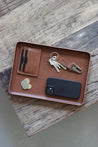 Eine Ablageschale aus Leder in Braun auf einer Holzplatte und in dieser liegt ein Handy, Kleingeld, ein Kartenetui und ein Schlüsselanhänger.