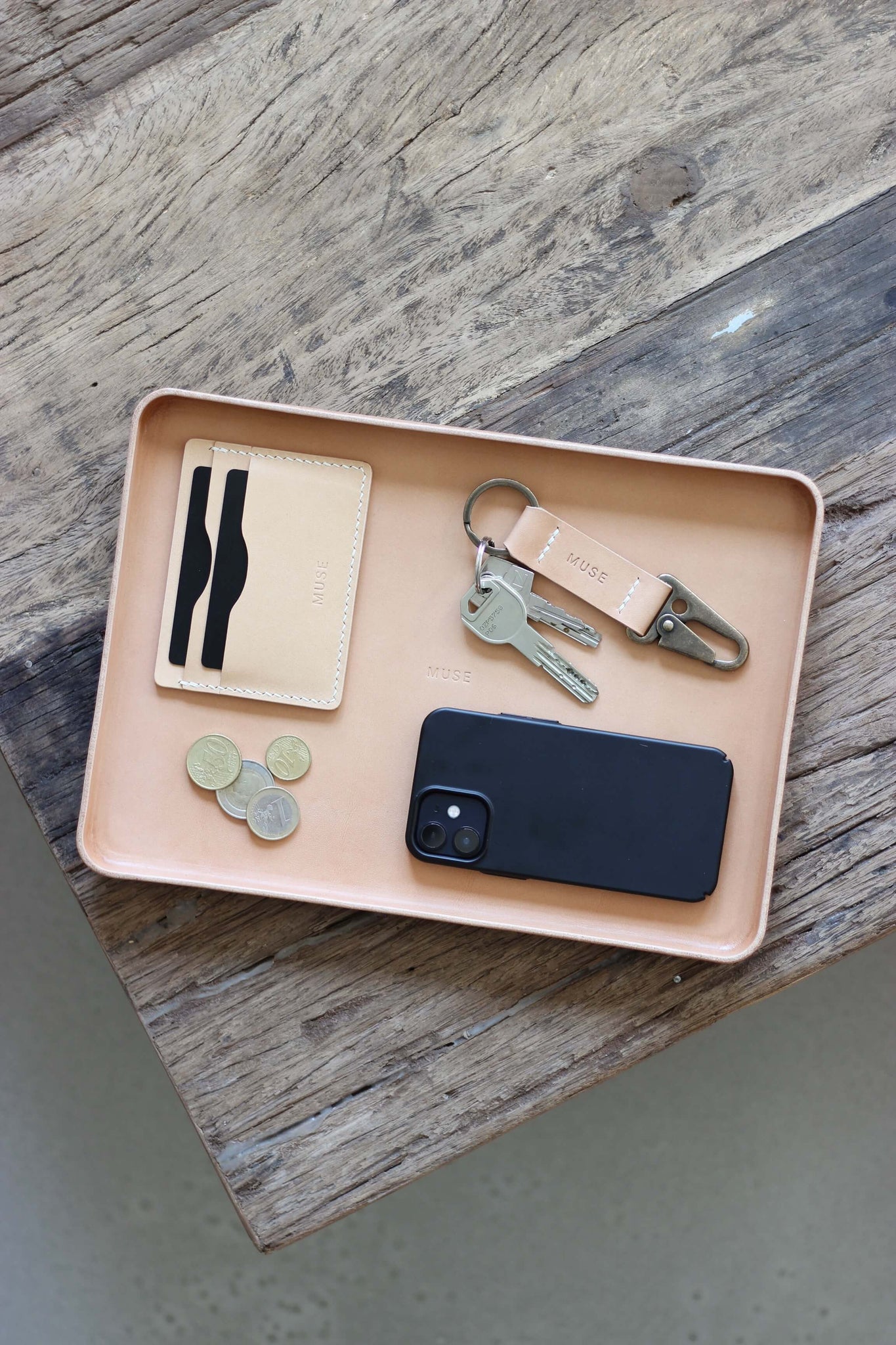 Eine Ablageschale aus Leder in Natural auf einer Holzplatte und in dieser liegt ein Handy, Kleingeld, ein Kartenetui und ein Schlüsselanhänger.