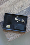 Eine Ablageschale aus Leder in Schwarz auf einer Holzplatte und in dieser liegt ein Handy, Kleingeld, ein Kartenetui und ein Schlüsselanhänger.