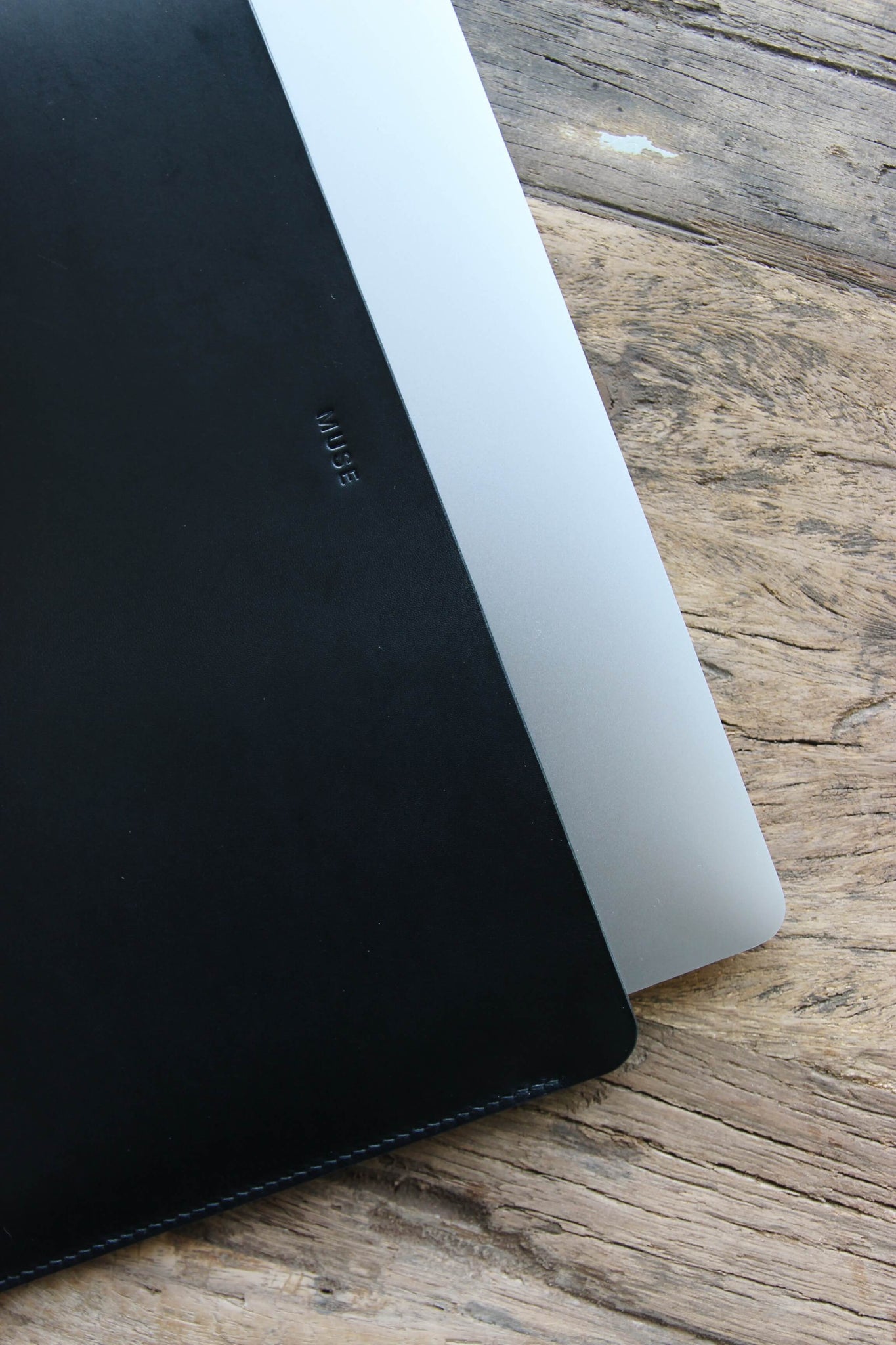 Auf einem Holztisch liegt eine Leder MacBook Hülle in Schwarz und aus dieser guckt ein kleines Stück eines MacBooks heraus.