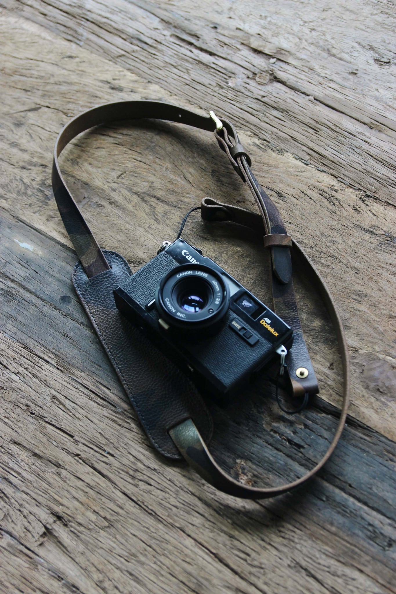 Eine Kamera mit einem Leder Kameragurt in Camo auf einem Holztisch liegend.