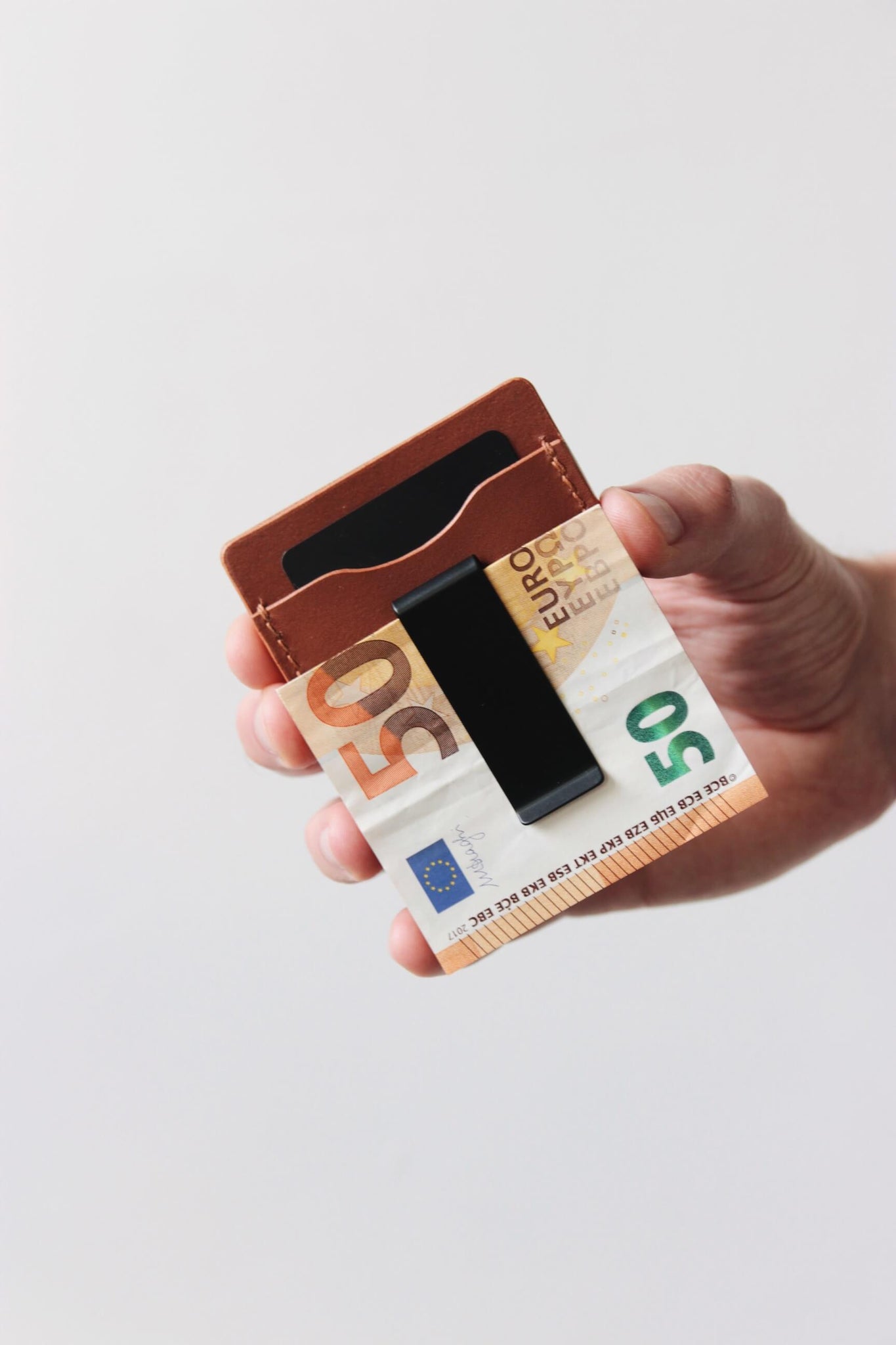 Eine Hand hält ein Kreditkartenetui aus Leder in Braun und in der integrierten Geldklammer steckt ein 50€-Schein.
