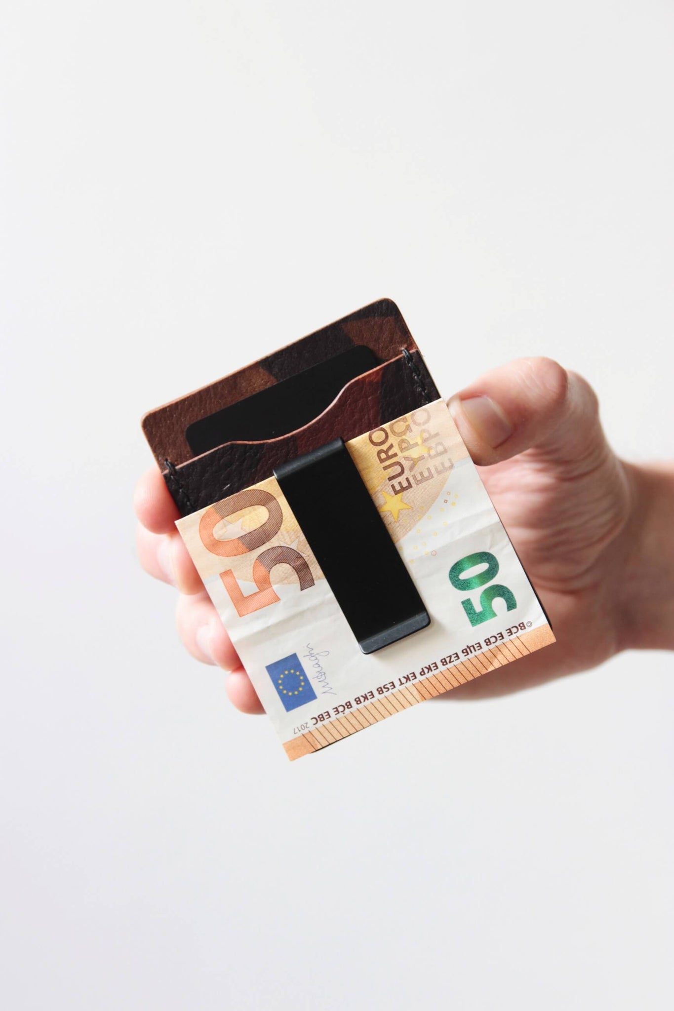 Eine Hand hält ein Kreditkartenetui aus Leder in Camouflage und in der integrierten Geldklammer steckt ein 50€-Schein.