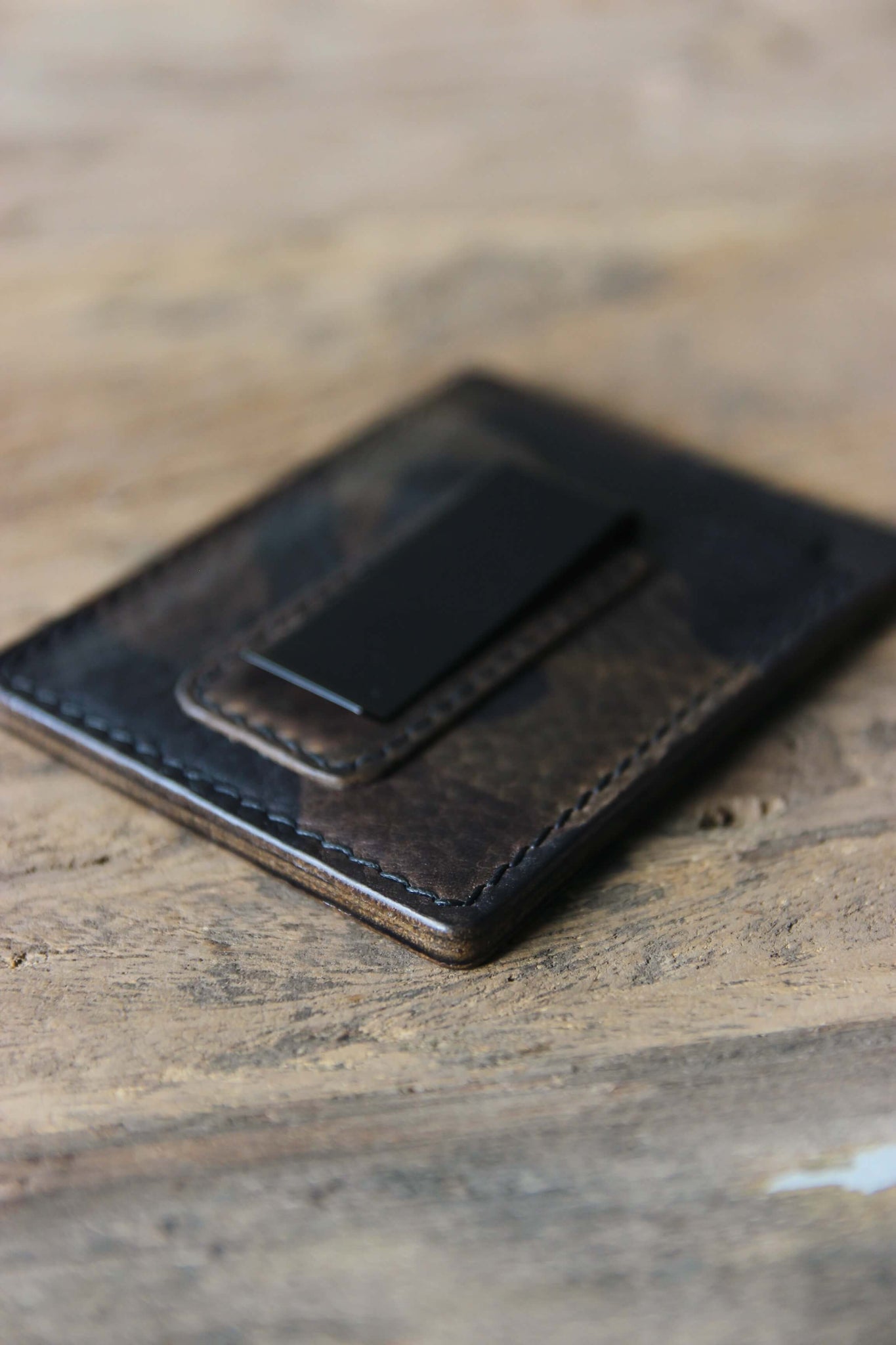 Nahaufnahme der schwarzen Geldklammer eines Kreditkartenetuis aus Leder in Camo auf einer Holzplatte liegend.