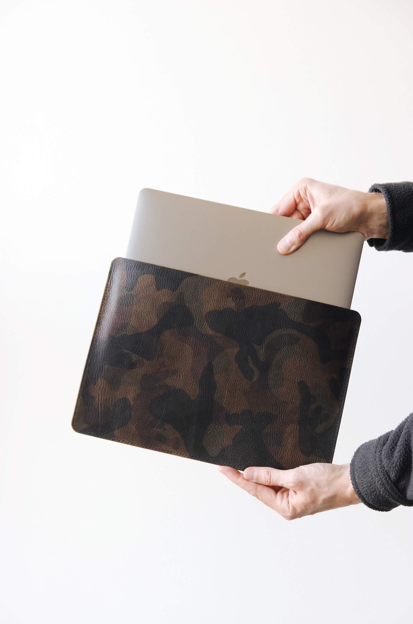 Eine Hand hält eine Leder MacBook Hülle in Camo und die andere Hand steckt ein MacBook in selbige.