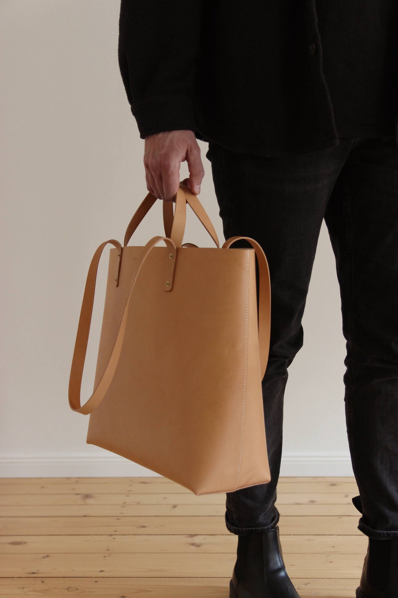 Ein Mann mit schwarzer Jeans und schwarzem Oberteil hält ein Leder Shopper in Natural an den kurzen Riemen in einer Hand.