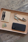 Detailaufnahme einer großen Ablageschale aus Leder in Natural auf einer Holzplatte und in dieser befindet sich ein Handy, Kleingeld, ein Kartenetui und ein Schlüsselanhänger.