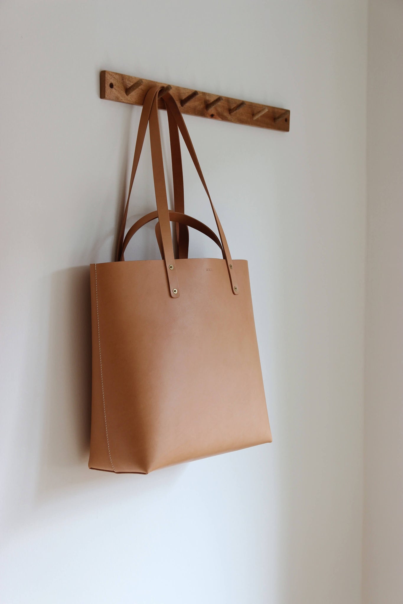 Ein Leder Shopper in Natural hängt an einer Garderobenleiste aus Holz.