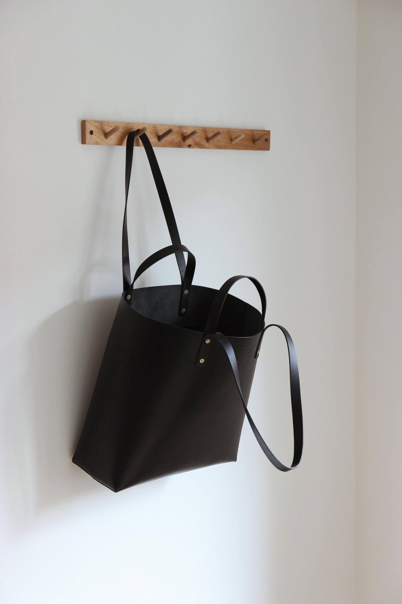 An einer Garderobenleiste aus Holz hängt an einem Henkel ein Leder Shopper in Schwarz.
