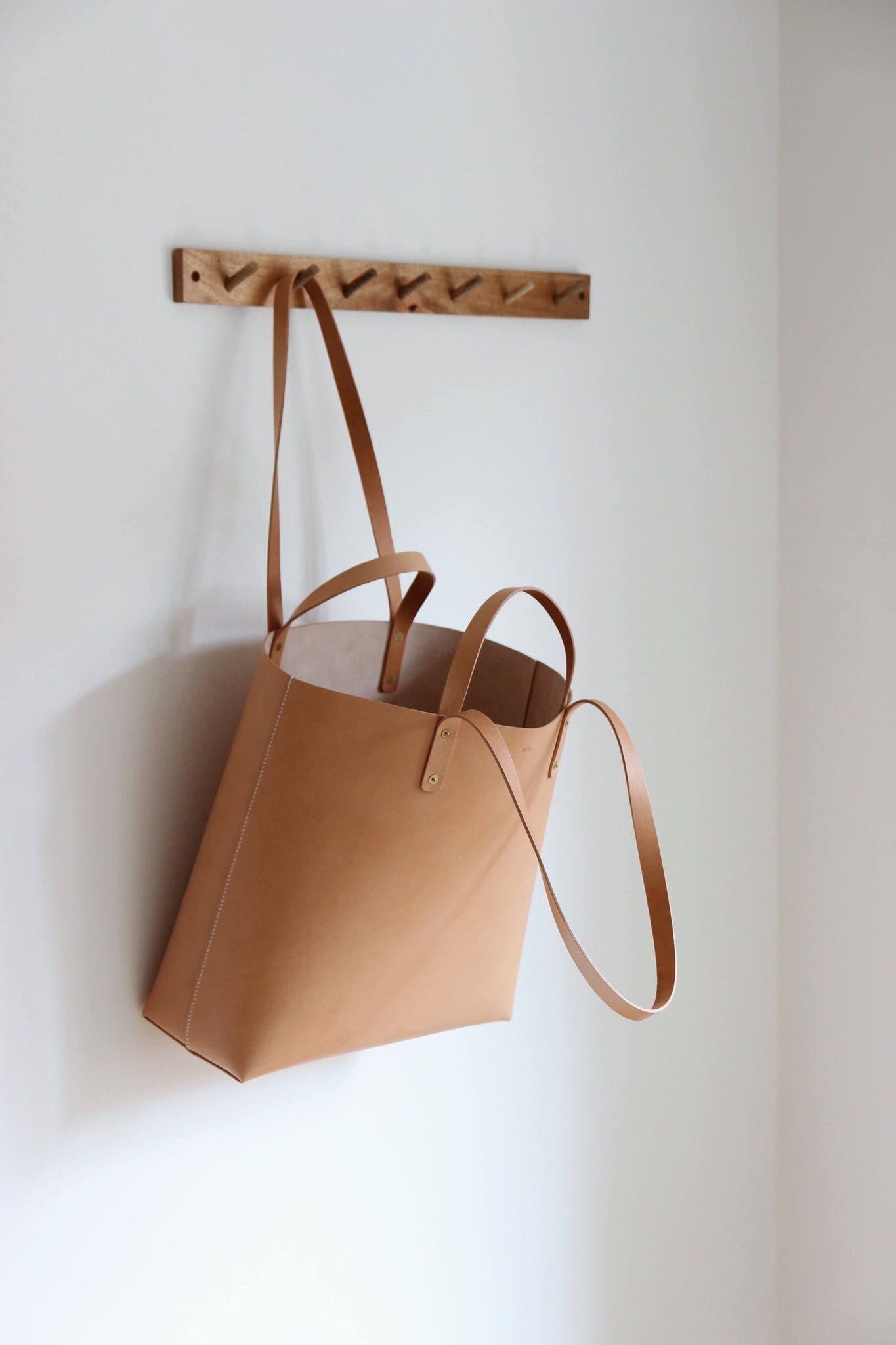 An einer Garderobenleiste aus Holz hängt an einem Henkel ein Leder Shopper in Natural.