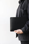 Eine Leder MacBook Hülle in Schwarz wird von jemandem in schwarzer Kleidung in der Hand gehalten.