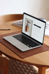 Ein aufgeklapptes MacBook liegt auf einer Schreibtischunterlage aus Leder in Braun auf einem Holztisch.