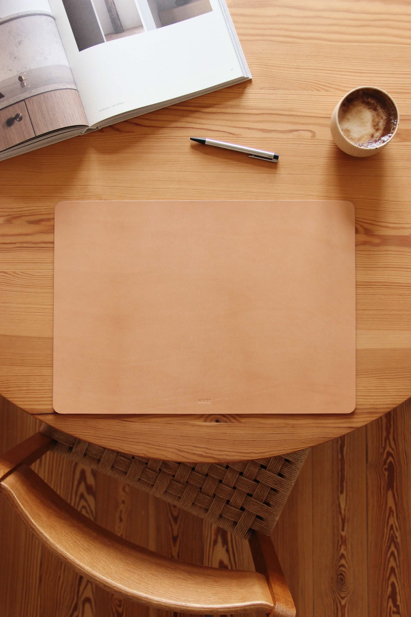 Auf einem Holztisch liegt eine Schreibtischunterlage aus Leder in Natural und als Dekoration eine Kaffeetasse, ein Stift und ein aufgeklapptes Buch.