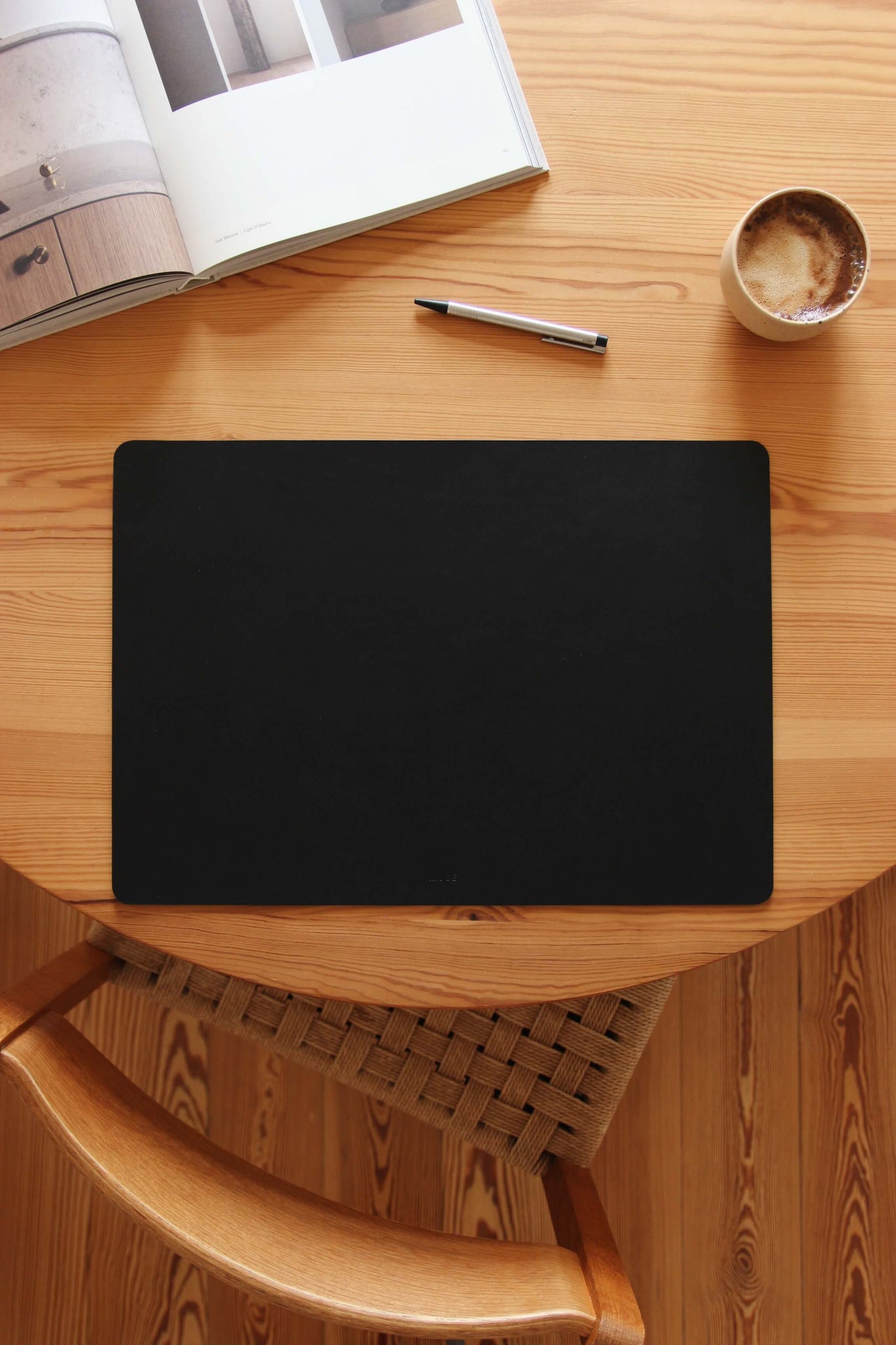 Auf einem Holztisch liegt eine Schreibtischunterlage aus Leder in Schwarz und als Dekoration eine Kaffeetasse, ein Stift und ein aufgeklapptes Buch.
