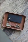 Eine Ablageschale aus Leder in Braun auf einer Holzplatte und in dieser befindet sich ein Handy und ein Schlüsselanhänger.