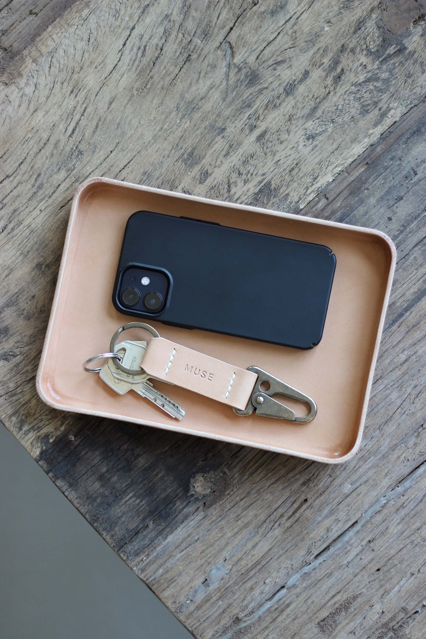 Eine Ablageschale aus Leder in Natural auf einer Holzplatte und in dieser befindet sich ein Handy und ein Schlüsselanhänger.