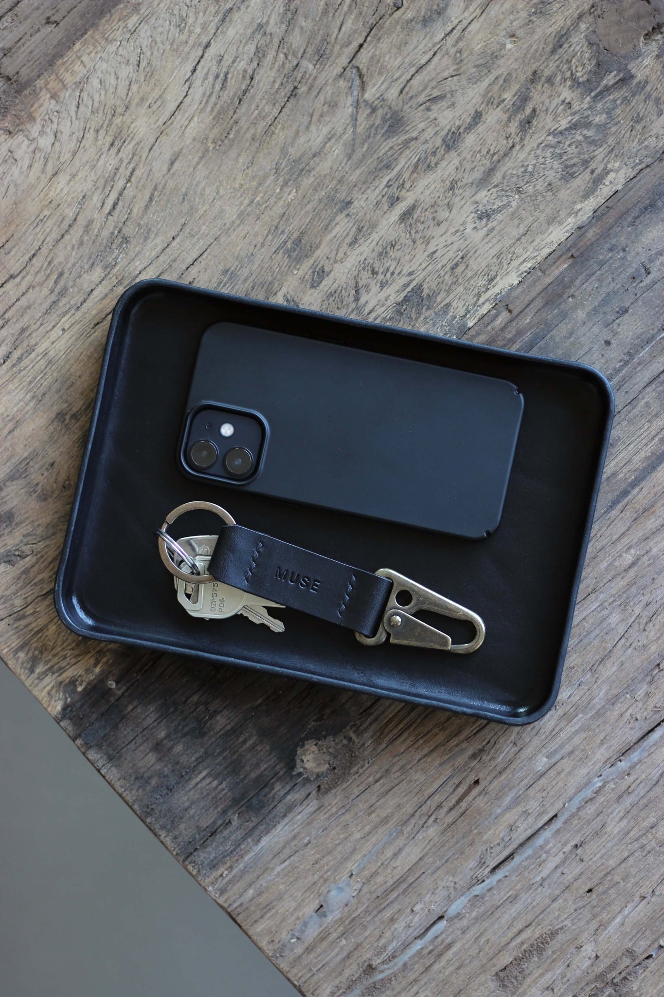 Eine Ablageschale aus Leder in Schwarz auf einer Holzplatte und in dieser befindet sich ein Handy und ein Schlüsselanhänger.