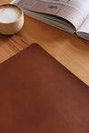 Lederkante einer Schreibtischunterlage aus Leder in Braun auf einer Holzplatte und eine Kaffeetasse und ein Buch.