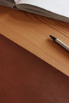 Ausschnitt einer Schreibtischunterlage aus Leder in Braun und ein Stift und ein aufgeklapptes Buch.