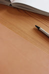 Ausschnitt einer Schreibtischunterlage aus Leder in Natural und ein Stift und ein aufgeklapptes Buch.