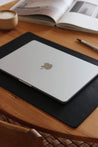 Seitenansicht einer Schreibtischunterlage aus Leder in Schwarz auf einem Holztisch und auf dieser befindet sich ein zugeklapptes MacBook.