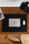 Leder Schreibtischunterlage in Schwarz auf einem Schreibtisch aus Holz und auf dieser befindet sich ein zugeklapptes MacBook und eine Maus.