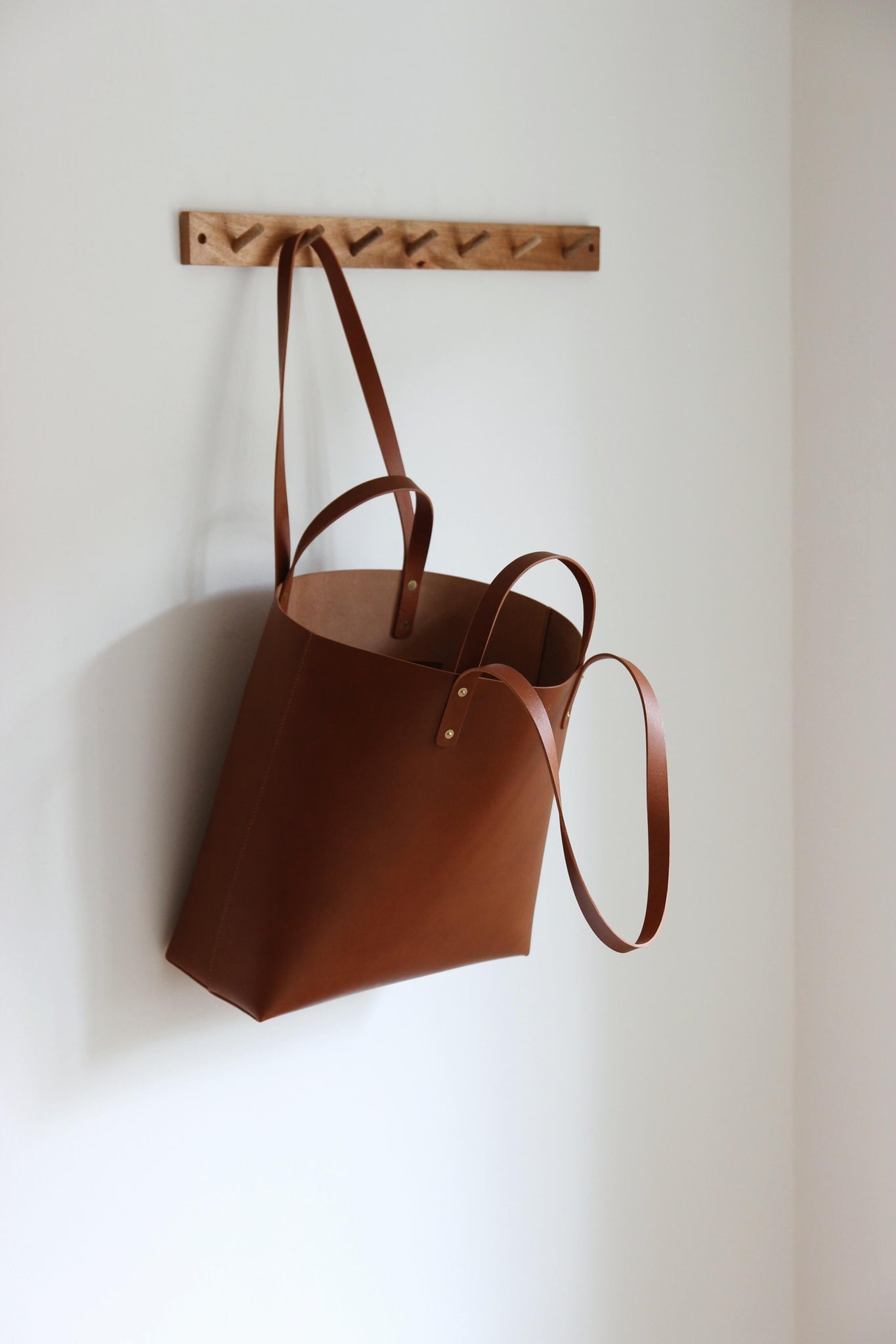 An einer Garderobenleiste aus Holz hängt an einem Henkel ein Leder Shopper in Braun.
